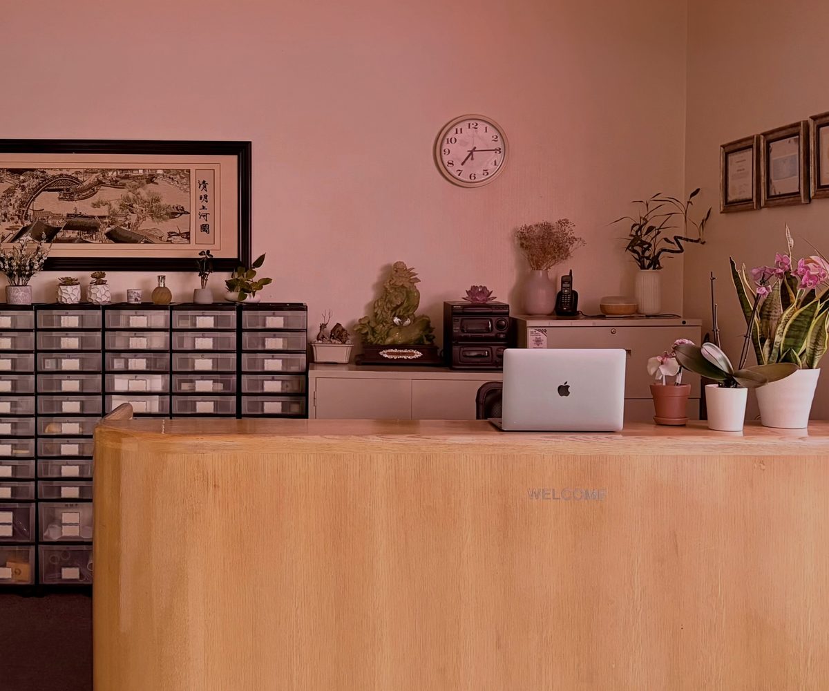 The A-Care reception desk: a light oak counter with orchids and tulips, a wall of labelled herb drawers behind it, and framed licences on the far wall.
